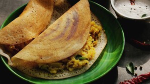 A plate with a folded crepe-like food item, possibly a crepe or pancake, filled with a yellow mixture, accompanied by a small bowl of white sauce and a red chili. - Deluxe Olive Residency (Kottayam)