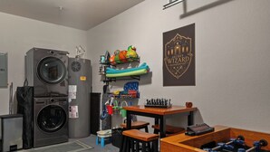 Ett tvättrum med tvättmaskin, torktumlare och strykbräda. Där finns också ett bord med ett fotbollsspel och en hylla med sportbollar.