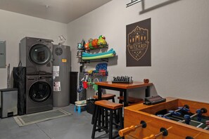 Ein Waschraum mit Waschmaschine, Trockner und Bügelbrett. Ein Tisch mit Tischfußball und ein Regal mit Sportbällen.