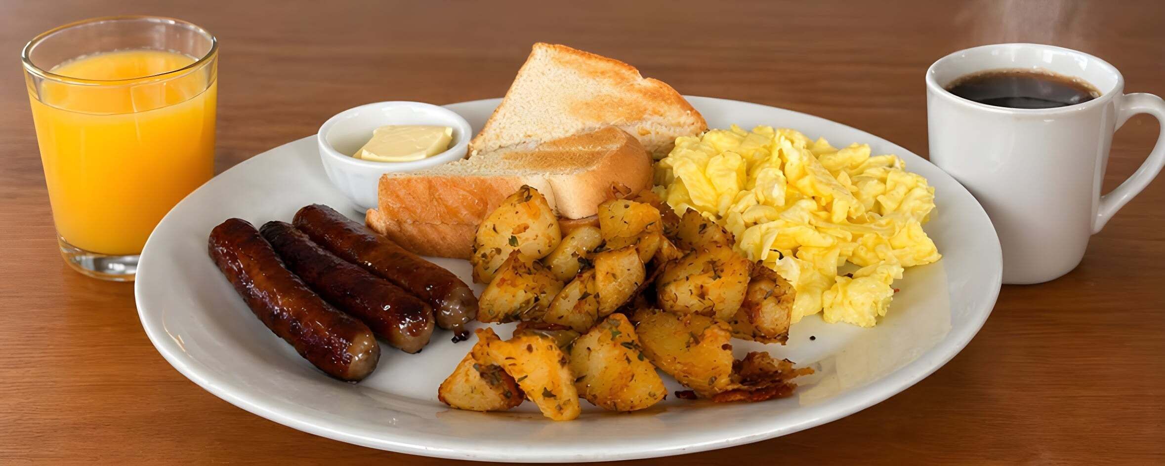Un plato de comida para el desayuno con salchichas, papas, huevos revueltos, tostadas, mantequilla y un vaso de jugo de naranja.