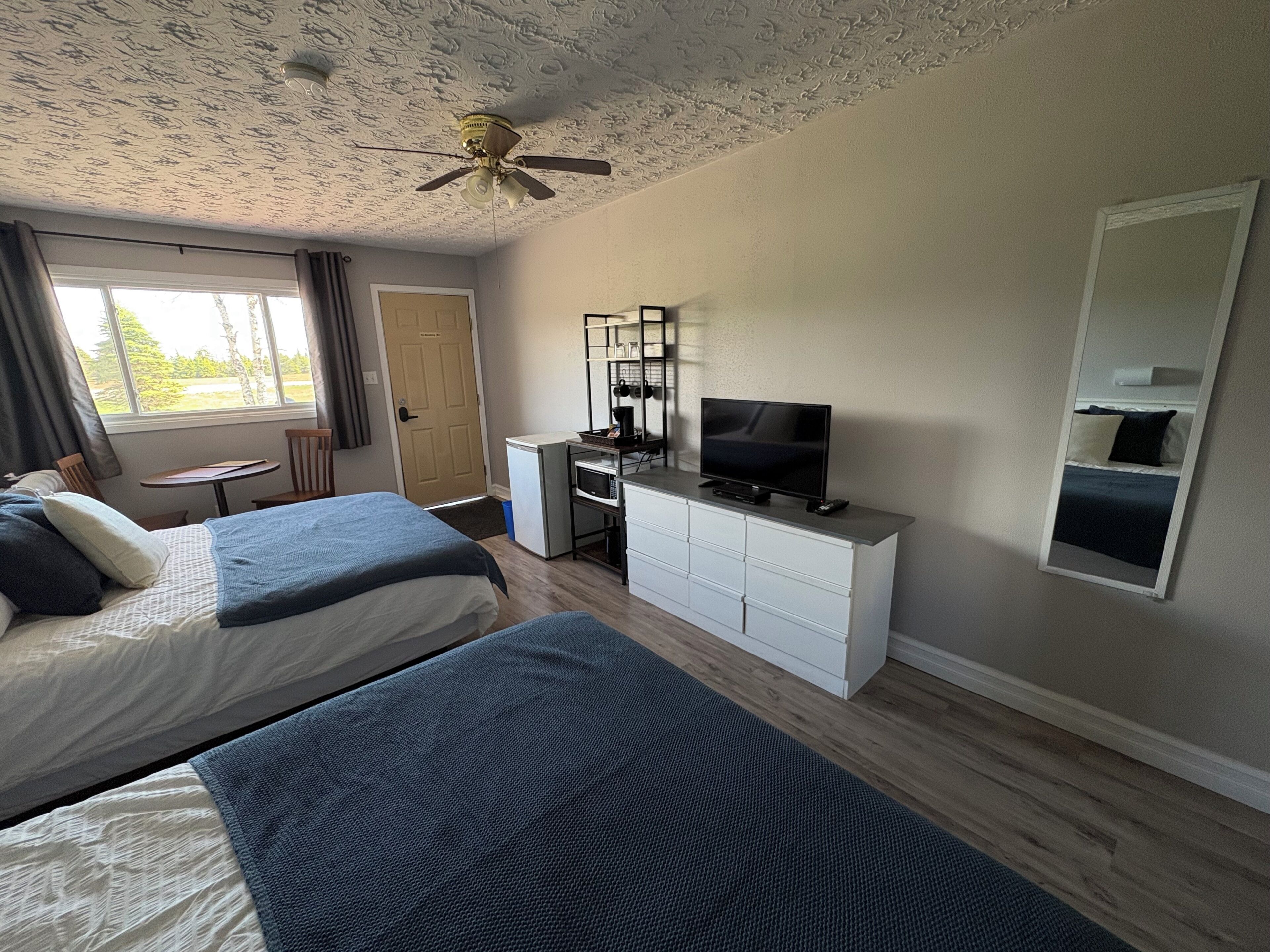 A hotel room with two beds, a desk with a TV, a ceiling fan, and a window with curtains.