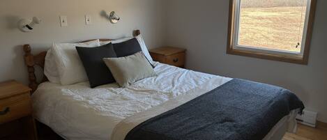 A neatly made bed with white bedding and a dark blue throw, a wooden nightstand with a lamp, and a window with blinds.