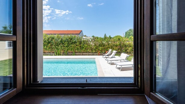 A view through a window of a swimming pool with lounge chairs, a clear sky, and a building in the background. - Villa del Rio by MMega (Strettoia)