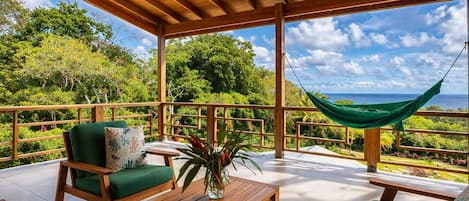 A covered outdoor seating area with wooden furniture, a hammock, and a view of the landscape.