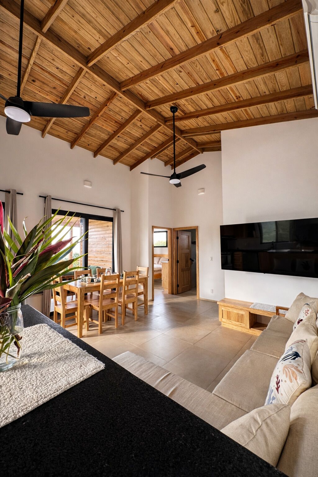 Een ruime woonkamer met een hoog houten plafond, een eethoek en een bank met decoratieve kussens.