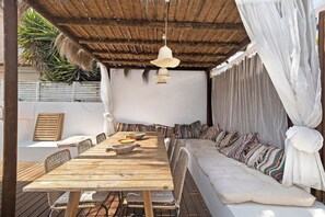 A wooden dining table with chairs, a sofa, and a hanging light fixture in a covered outdoor area.