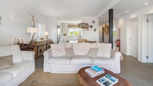 A modern living room with a white sofa, a wooden coffee table, and a dining area with a wooden cabinet and a hanging light fixture.
