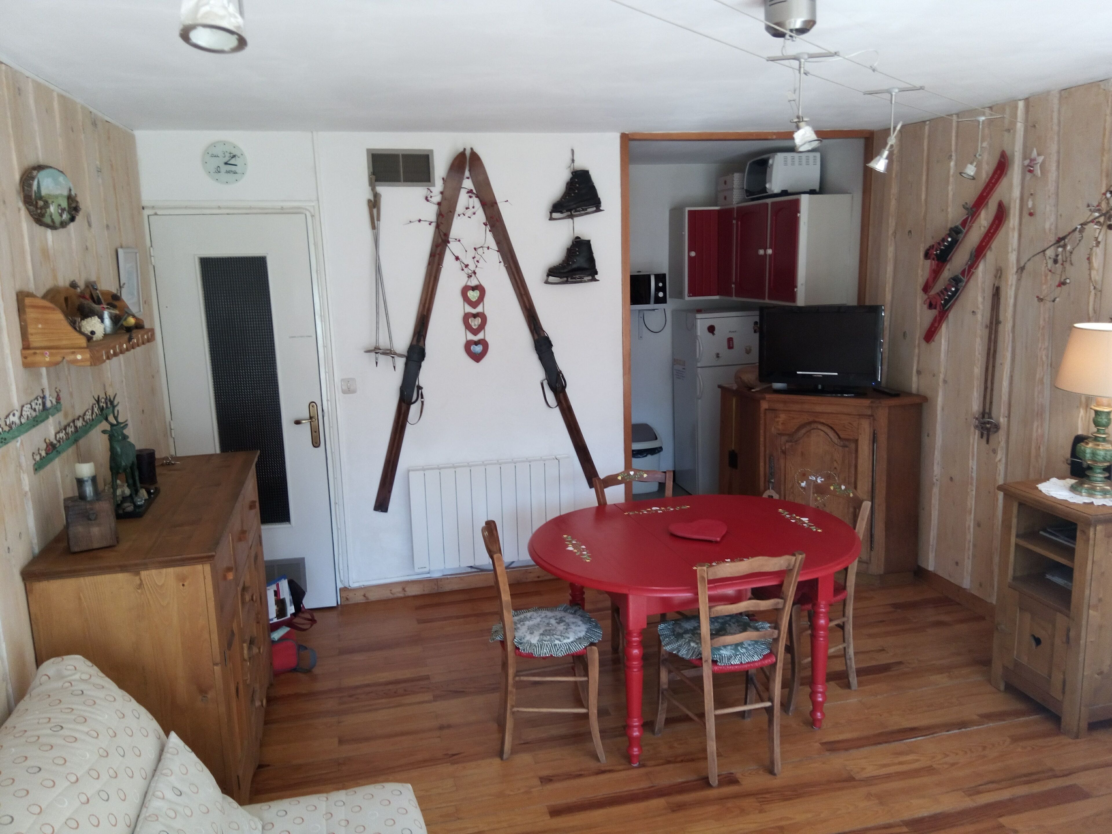Una habitación con piso de madera, una mesa de comedor roja con cuatro sillas, un aparador de madera, un televisor sobre un mueble de madera y esquíes colgados en la pared.
