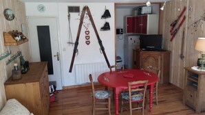 A room with a wooden floor, a red dining table with four chairs, a wooden cabinet, a television on a wooden stand, and skis hanging on the wall. - STOP: My spacious two-bedroom apartment across from Écrins Park (Orcières)