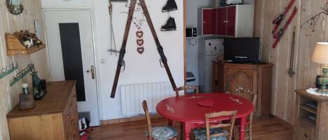 Una habitación con piso de madera, una mesa de comedor roja con cuatro sillas, un aparador de madera, un televisor sobre un mueble de madera y esquíes colgados en la pared.