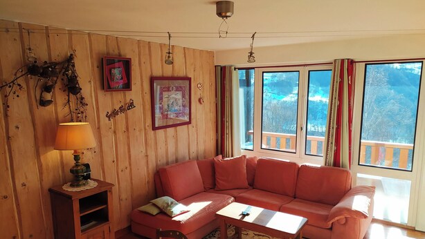 A wooden-paneled room with a red sofa, a small table, and a lamp. There are two windows with curtains, and a picture frame on the wall. - STOP: My spacious two-bedroom apartment across from Écrins Park (Orcières)
