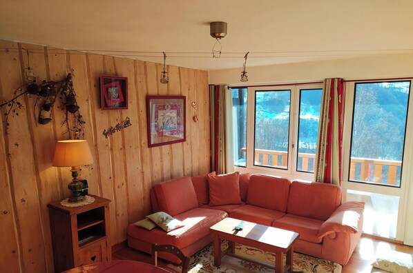 Habitación con paneles de madera, un sofá rojo, una mesita y una lámpara. Cuenta con dos ventanas con cortinas y un cuadro colgado en la pared.