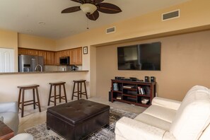 A living room with a sofa, ottoman, and bar stools. There is a kitchen area with cabinets and appliances. A ceiling fan and a flat-screen TV are visible. - Alii Cove II-4 | Monthly Rental in Kona | 30-Day Stay Alii Drive (Kailua Kona)