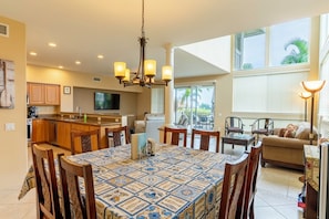 A spacious kitchen with wooden cabinets, a dining area with a patterned tablecloth, and a living area with a sofa and chairs. - Alii Cove II-4 | Monthly Rental in Kona | 30-Day Stay Alii Drive (Kailua Kona)