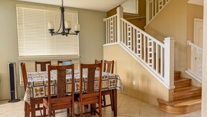 A dining area with a table set for four, a staircase with white railings, and a chandelier. - Alii Cove II-4 | Monthly Rental in Kona | 30-Day Stay Alii Drive (Kailua Kona)