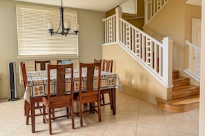 A dining area with a table set for four, a staircase with white railings, and a chandelier. - Alii Cove II-4 | Monthly Rental in Kona | 30-Day Stay Alii Drive (Kailua Kona)