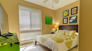 A hotel room with a bed, a desk, a window with blinds, a wall-mounted lamp, and artwork. - Alii Cove II-4 | Monthly Rental in Kona | 30-Day Stay Alii Drive (Kailua Kona)
