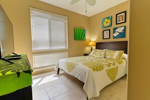 A hotel room with a bed, a desk, a window with blinds, a wall-mounted lamp, and artwork. - Alii Cove II-4 | Monthly Rental in Kona | 30-Day Stay Alii Drive (Kailua Kona)