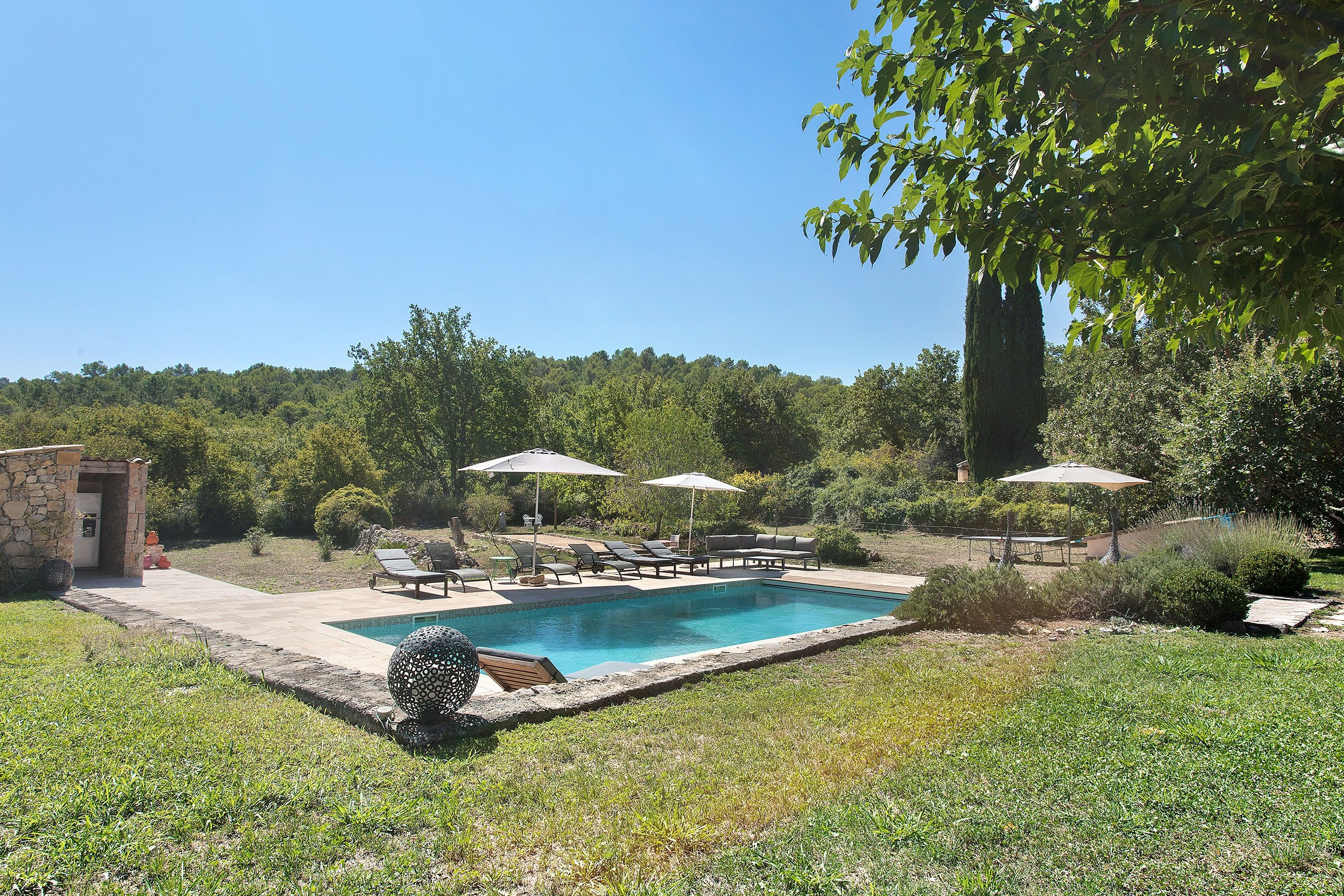 Une piscine entourée de verdure et d’un bâtiment en pierre, avec des chaises longues et des parasols à proximité.