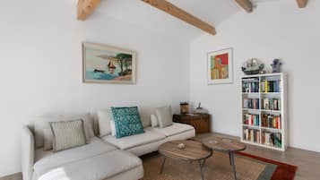 Een woonkamer met een witte bank, twee salontafels, een boekenkast en twee kunstwerken in lijsten aan de muur.