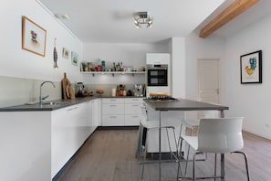 A modern kitchen with white cabinetry, a central island with bar stools, and a built-in oven. - Villa 'Maison Mennoise' with Swimming Pool (Tourrettes)