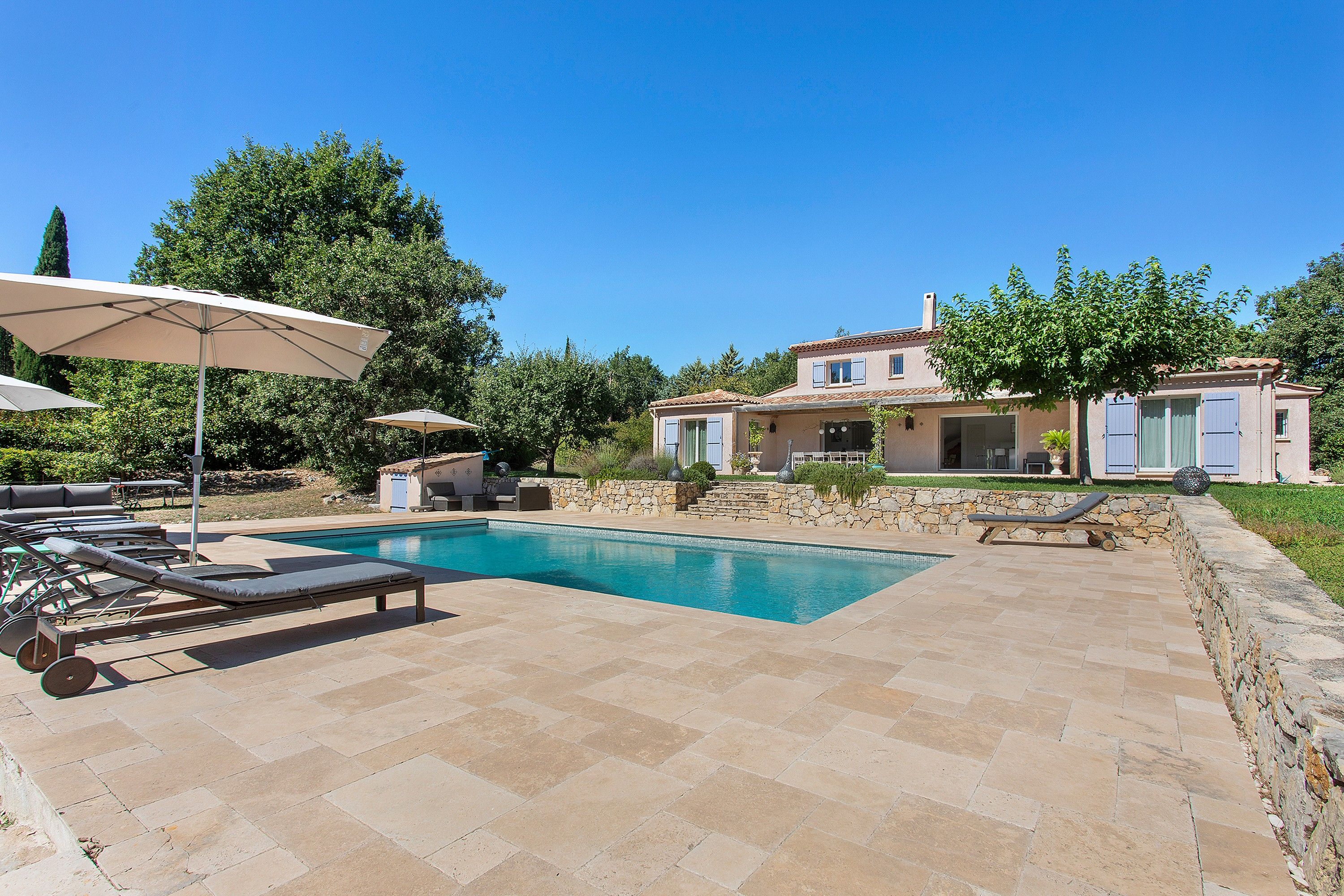 A pool area with lounge chairs and umbrellas, a stone patio, and a house with a tiled roof.