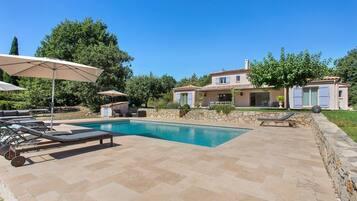 A pool area with lounge chairs and umbrellas, a stone patio, and a house with a tiled roof.