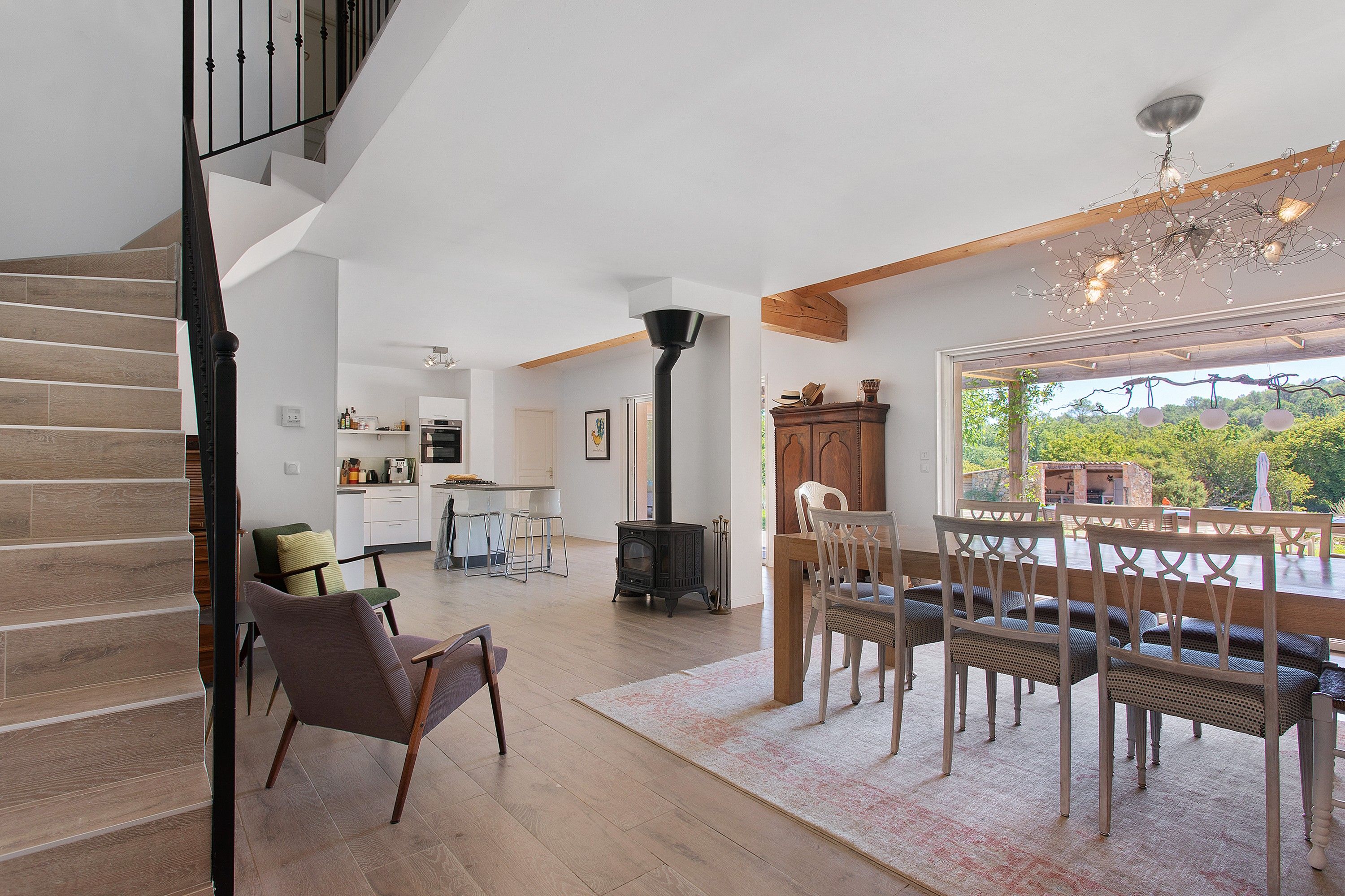 A spacious living area with a staircase, a dining table with chairs, a kitchen area, and a view of the outdoors.