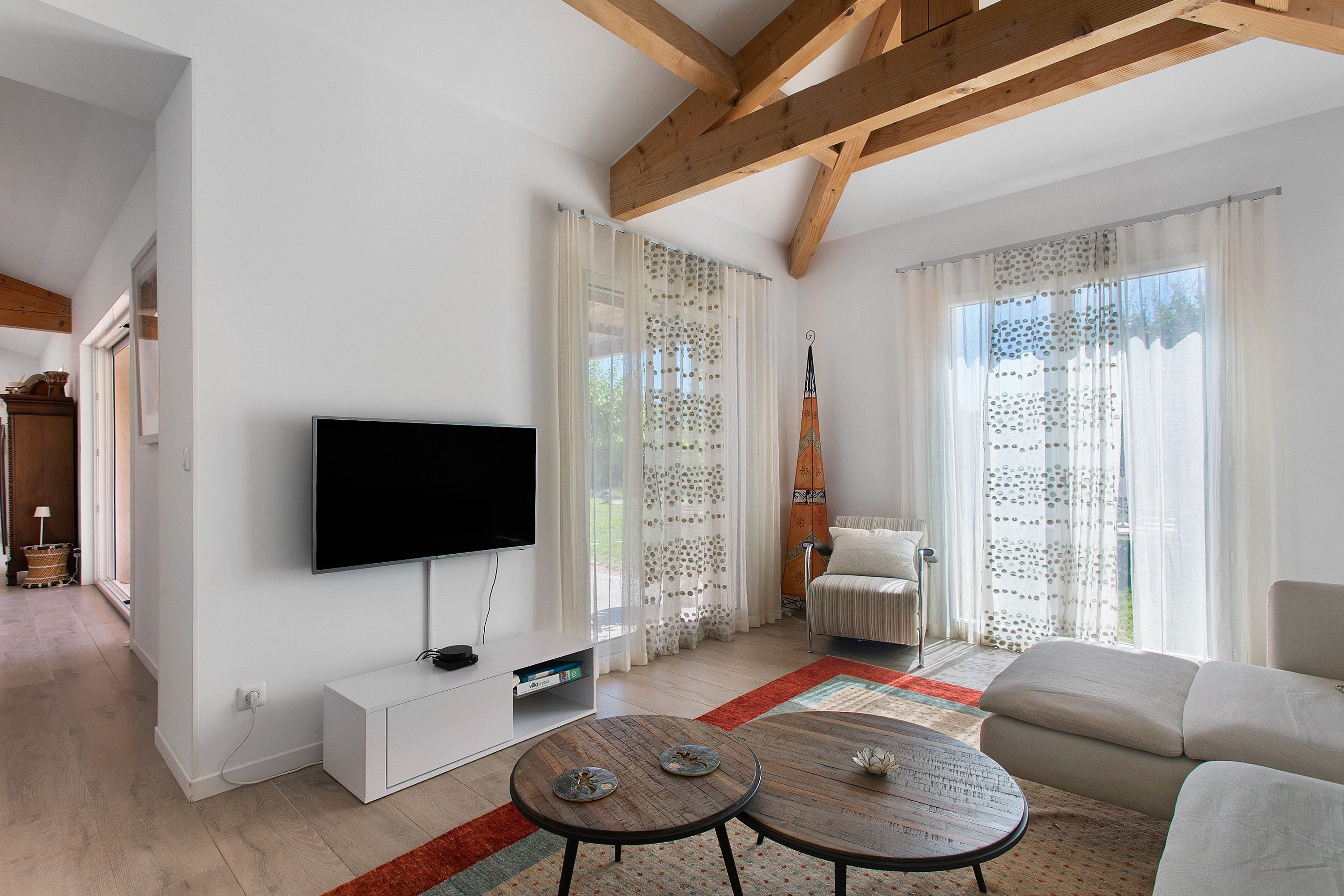 Un salon moderne doté d’un téléviseur à écran plat, d’un meuble de rangement blanc, de deux petites tables rondes, d’un canapé et d’une grande fenêtre agrémentée de rideaux à motifs.