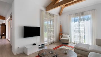 Un salon moderne doté d’un téléviseur à écran plat, d’un meuble de rangement blanc, de deux petites tables rondes, d’un canapé et d’une grande fenêtre agrémentée de rideaux à motifs.