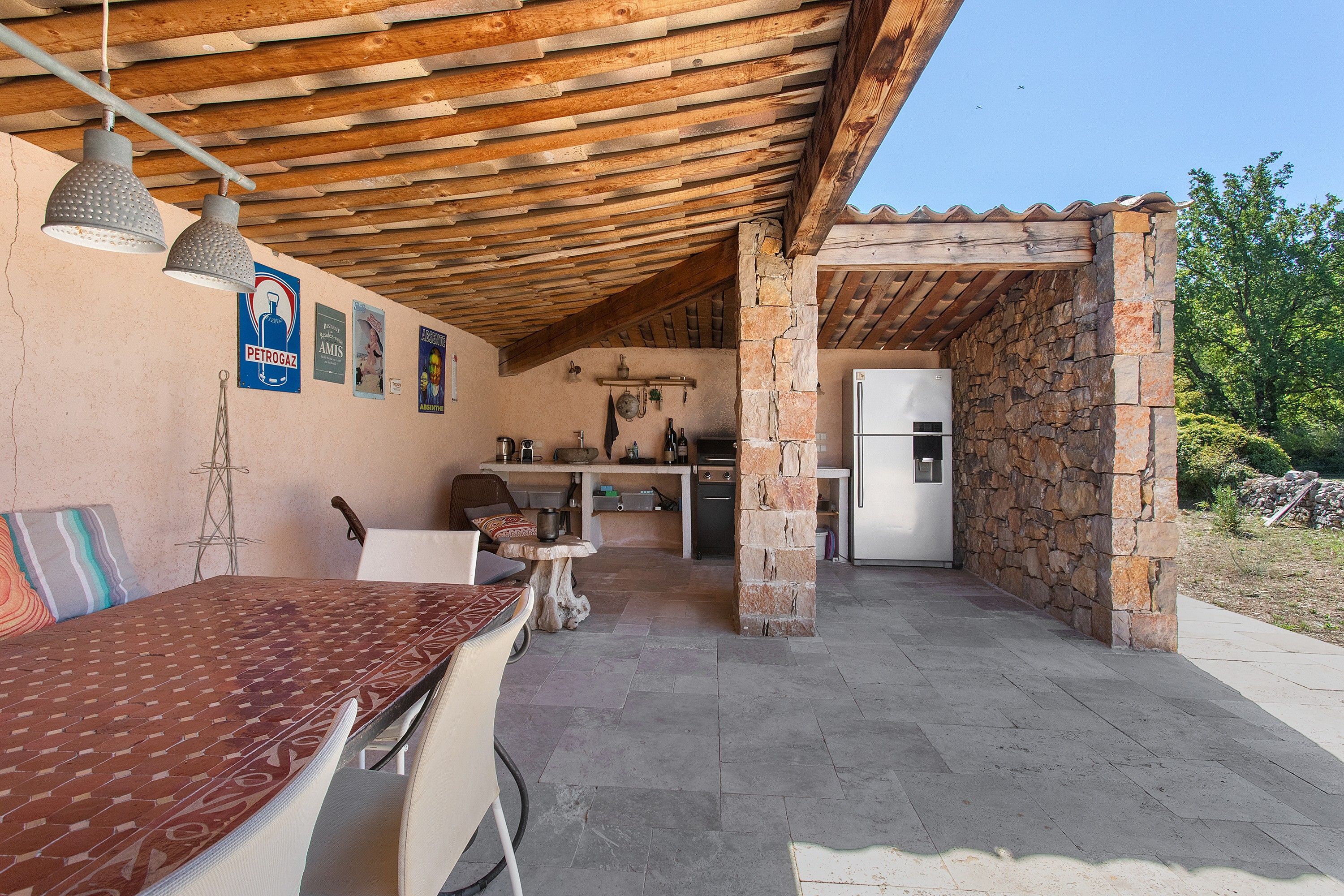 Un espace extérieur abrité, aménagé pour les repas, avec une table et des chaises, un coin cuisine équipé d’un réfrigérateur, et des murs en pierre.