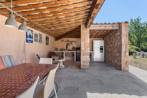 A covered outdoor dining area with a table and chairs, a kitchen area with a refrigerator, and stone walls. - Villa 'Maison Mennoise' with Swimming Pool (Tourrettes)