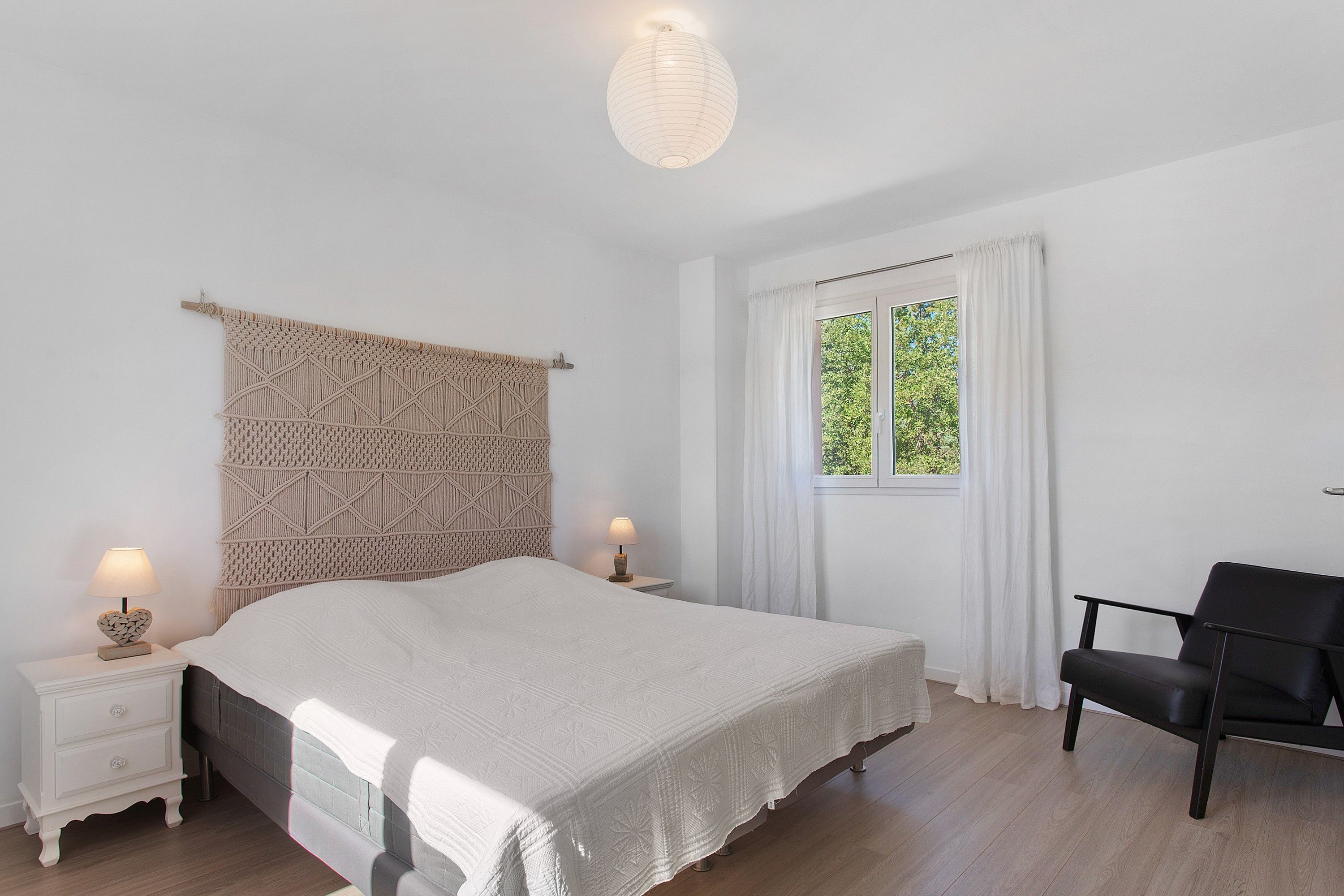 A bedroom with a bed, two bedside tables, a chair, a window with curtains, and a hanging light fixture.