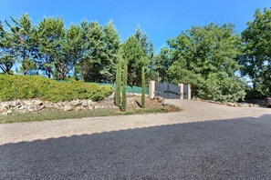 Ein Schotterweg führt zu einem verschlossenen Eingang mit einem Gebäude, umgeben von Bäumen und einer Hecke.