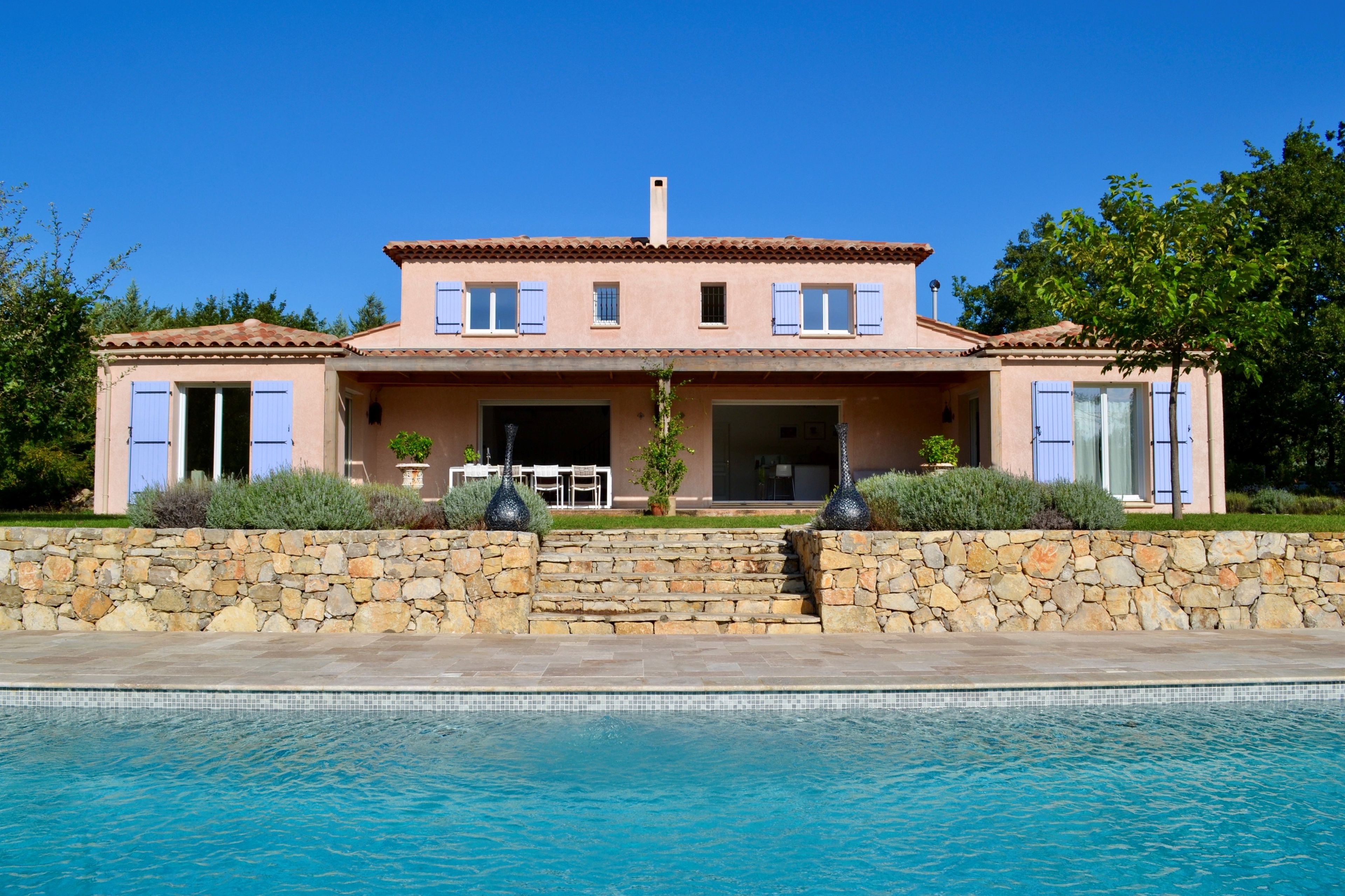 Une maison à deux étages avec un mur de pierre à l’avant, une piscine et un ciel d’un bleu limpide.