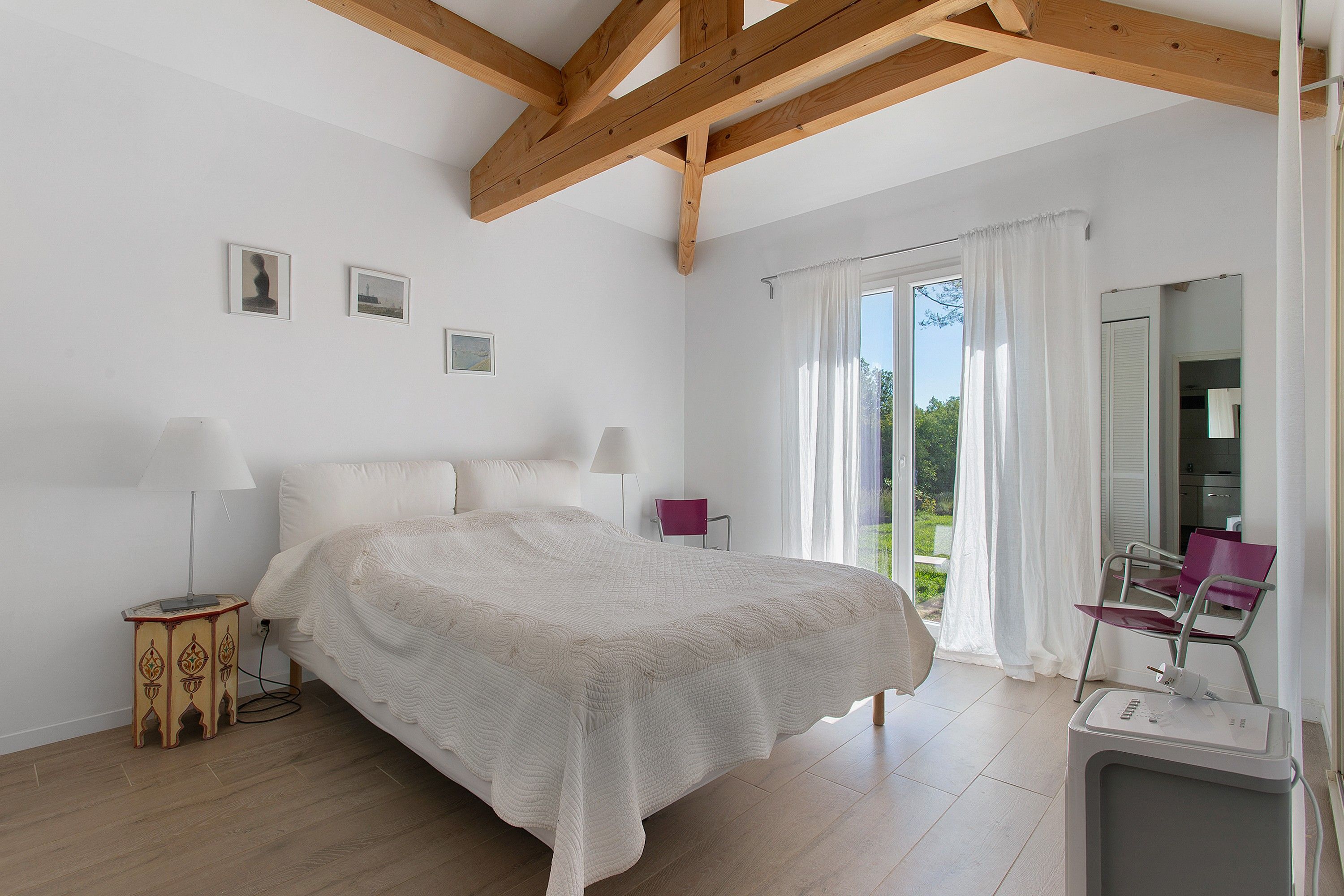 A bedroom with a large bed, a wooden ceiling, a window with curtains, and a small table with a lamp.