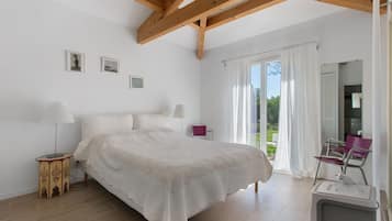 A bedroom with a large bed, a wooden ceiling, a window with curtains, and a small table with a lamp.