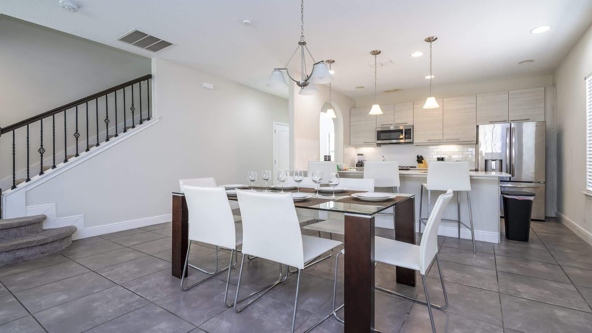 Cozinha moderna com eletrodomésticos de aço inoxidável, área de jantar com mesa de vidro e cadeiras brancas, e escada com corrimão de metal.