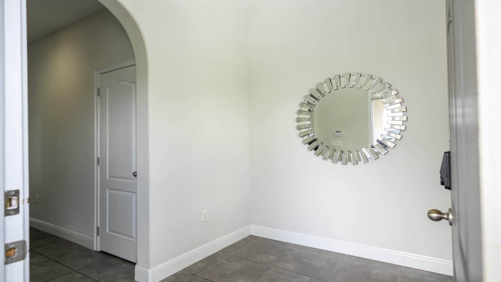 Un couloir avec une porte blanche, un miroir rond accroché au mur et un sol carrelé de couleur grise.