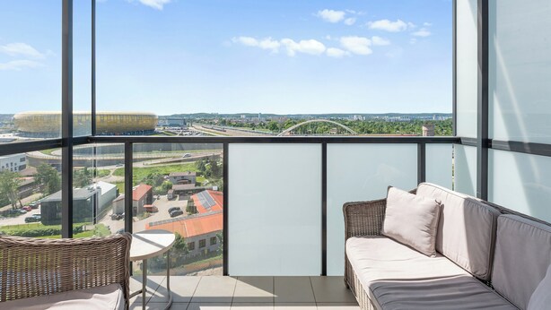 A balcony with a wicker sofa, a small table, and a glass wall offering a city view. - RentPlanet - Apartament Sucha (Gdańsk)