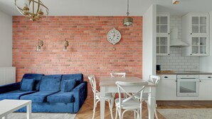 A living room with a blue sofa, white dining table, and chairs, a brick wall, and a kitchen area with white cabinets and an oven. - RentPlanet - Apartament Sucha (Gdańsk)