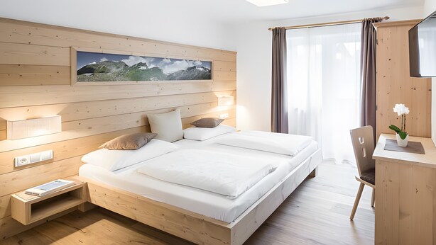 Una cama de madera con un cuadro colgado, lámparas de noche, un escritorio con una silla y una ventana con cortinas.