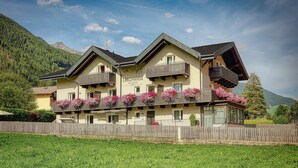 Un hotel de varios pisos con balcones decorados con flores, una valla de madera al frente y montañas al fondo.