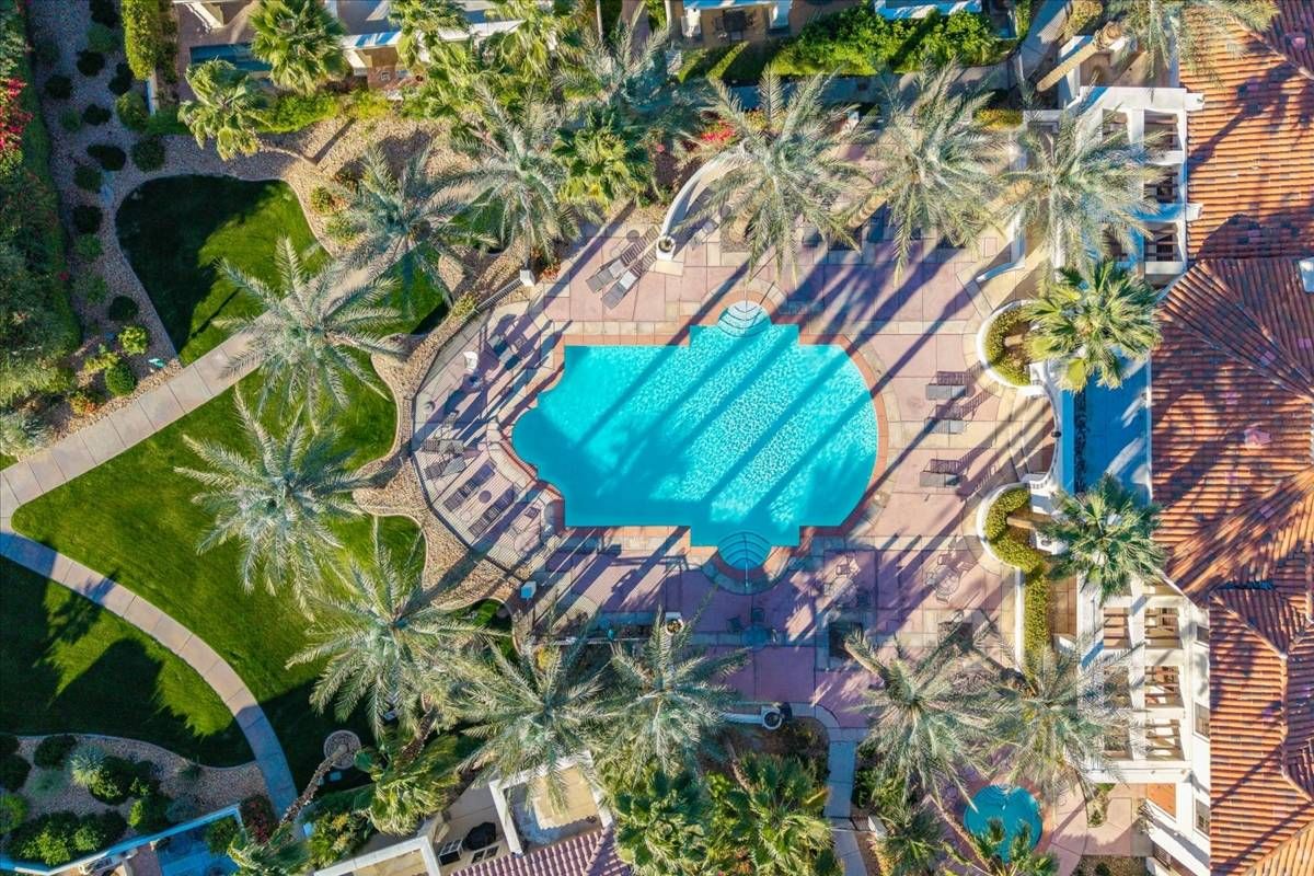 An aerial view of a resort with a central swimming pool surrounded by palm trees and a tiled roof.