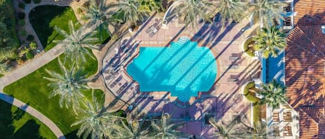 An aerial view of a resort with a central swimming pool surrounded by palm trees and a tiled roof.