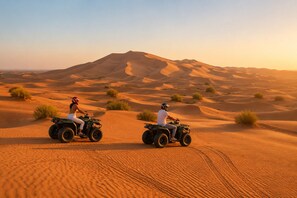 Duas pessoas pilotando veículos todo-terreno em uma duna do deserto, com uma montanha ao fundo.