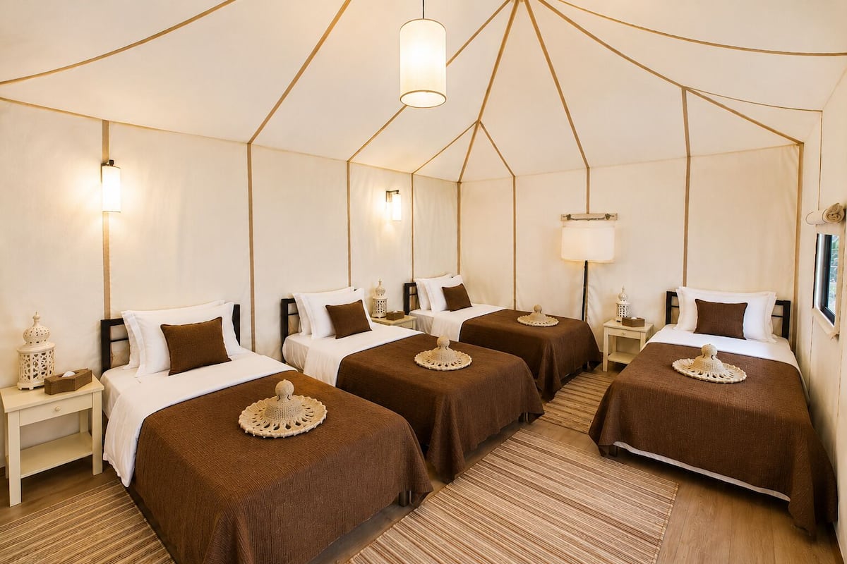 Habitación de hotel con tres camas, cada una con una colcha marrón y un adorno decorativo al pie. Hay mesitas de noche con lámparas, un espejo grande y una ventana.