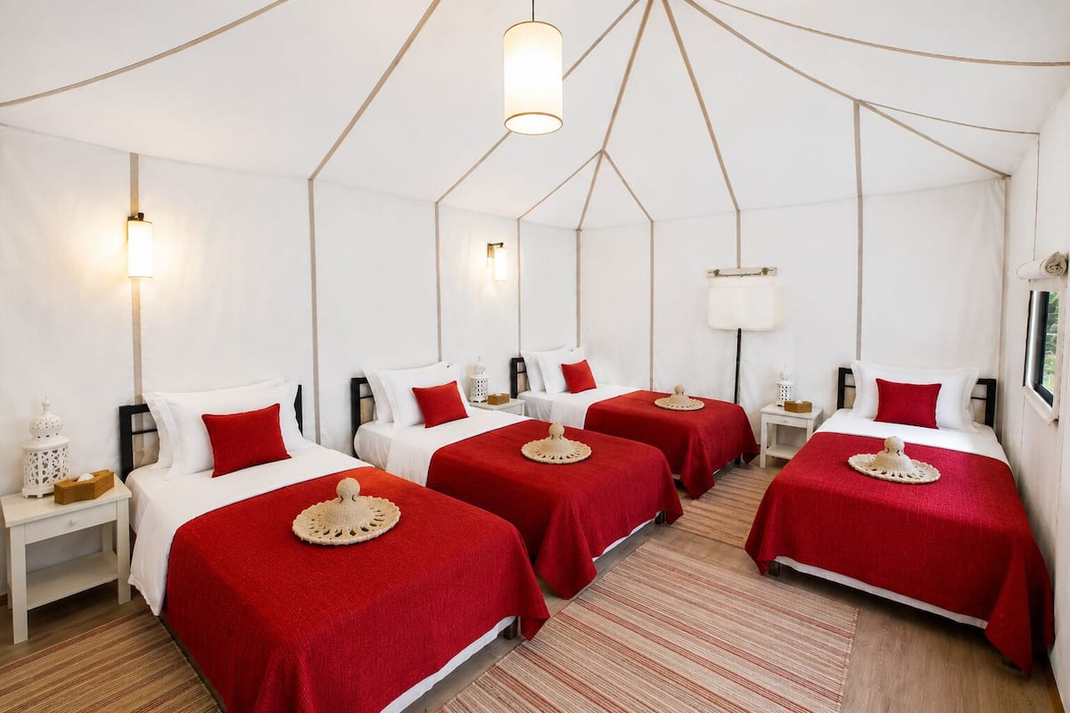 Habitación de hotel con cuatro camas, cada una con una colcha roja y un adorno en la cama. Hay mesitas de noche blancas con lámparas, una pared blanca y un techo con diseño geométrico.