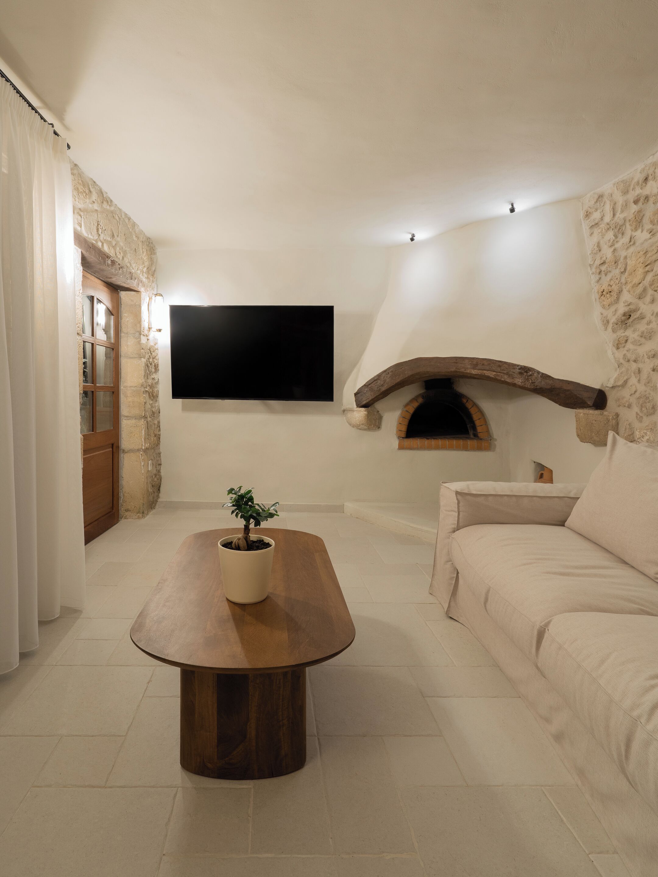 A living room with a stone wall, a flat-screen TV, a wooden coffee table with a potted plant, a sofa, and a stone fireplace.