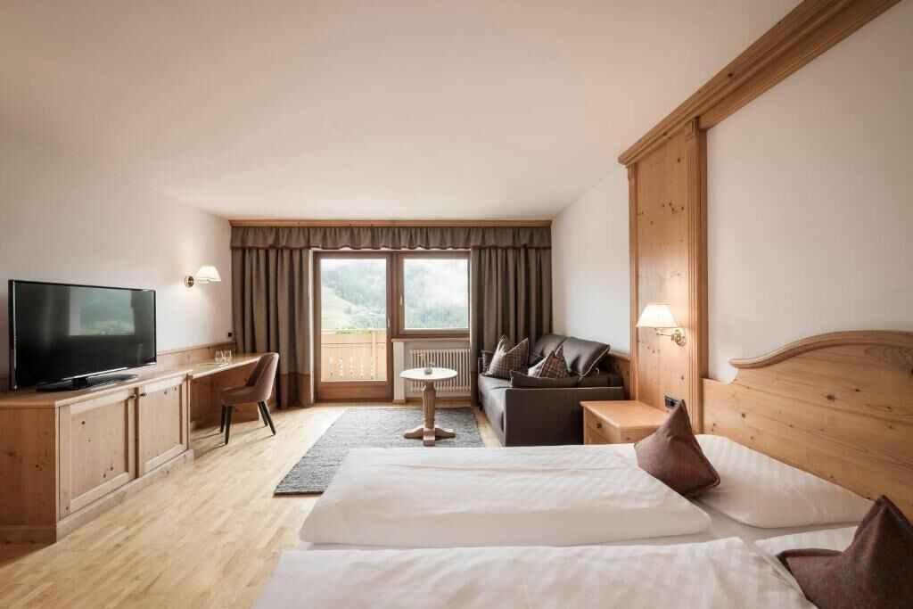 Habitación de hotel con cama, televisor, escritorio, silla, sofá y ventana con cortinas.